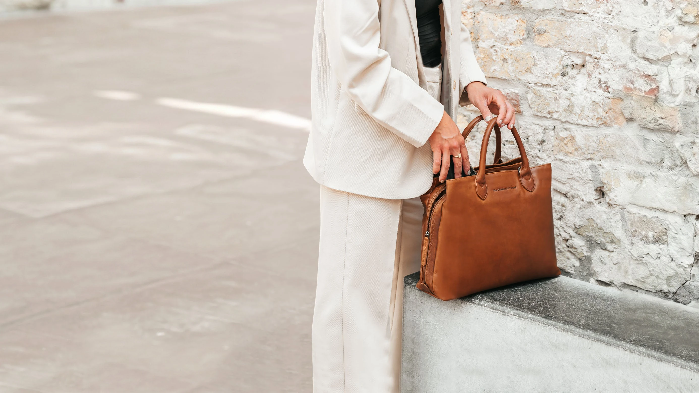 Woman in a light suit holding a cognac Chesterfield leather bag beside a stone wall
